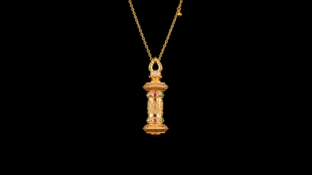 Gold-tone prayer-wheel-inspired pendant with a cylindrical relief body, colored accents and an open teardrop bail on a black background.