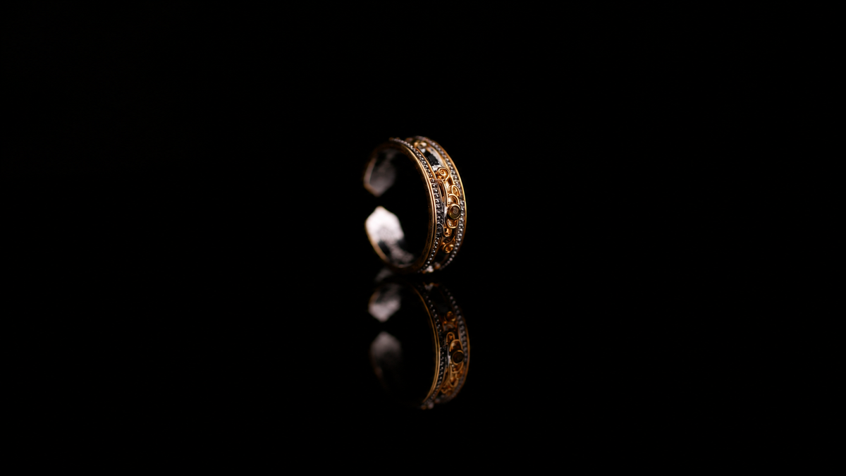 Open ring with gold-tone spiral scrolls, beaded borders, a central amber-toned detail, and reflection against black.