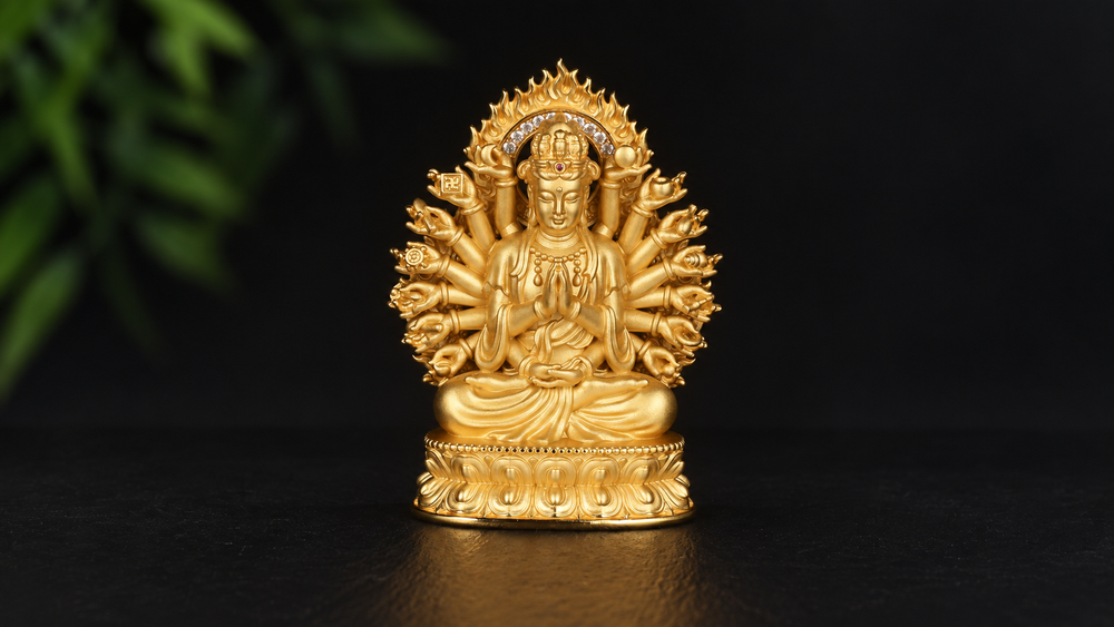 Gold-tone Thousand-Armed Guanyin sculpture with central prayer hands, a fan of arms, multiple heads, flame-shaped aureole, and a layered lotus base against a black background.